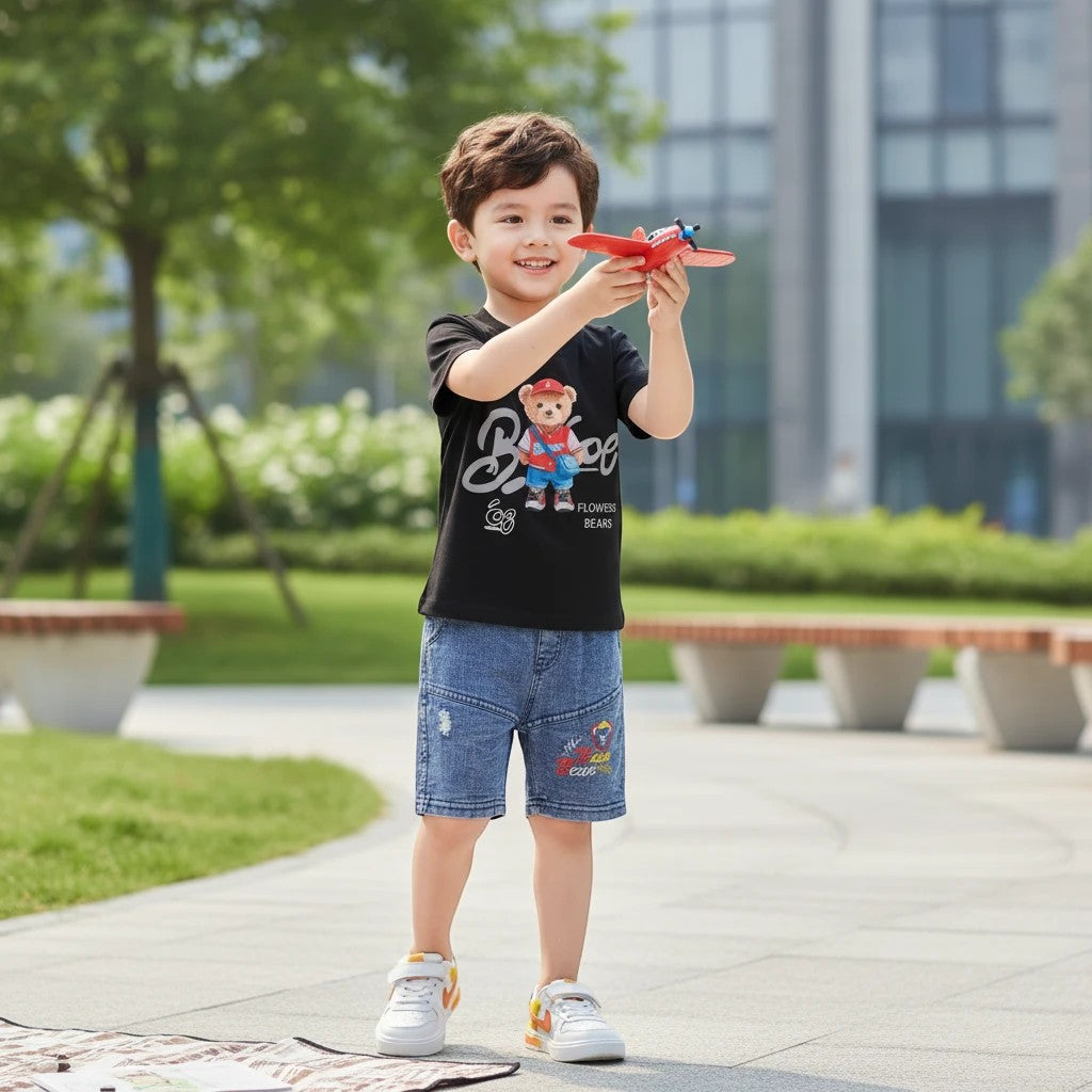 Mini & Mama black cotton t-shirt with bear graphic and text, paired with distressed denim shorts. Boy playing with toy airplane.