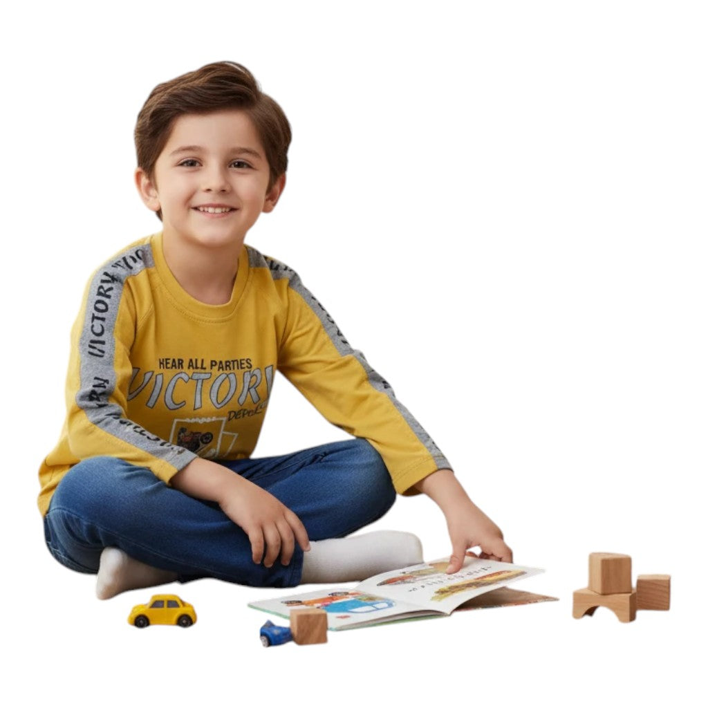 Mini & Mama boys yellow long sleeve shirt with "Victory" graphic and grey sleeves, paired with blue jeans. Boy sitting and reading.